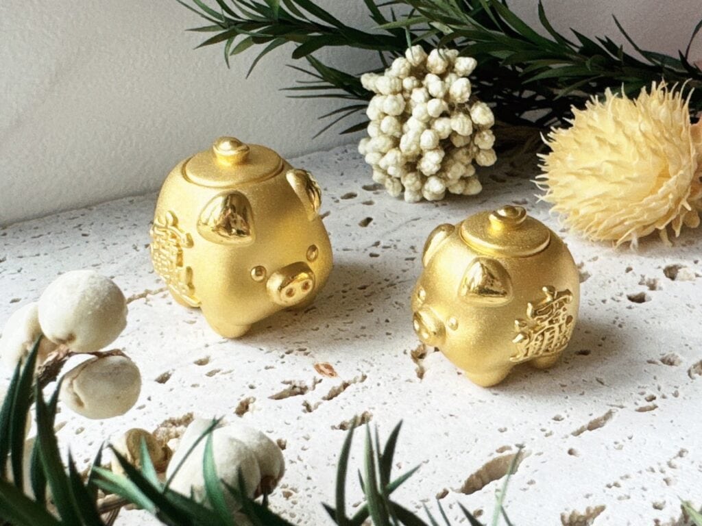 兩隻金色小豬造型擺飾，表面刻有招財字樣，擺放於白色石紋桌面與乾燥花裝飾之間，象徵財運與好運。