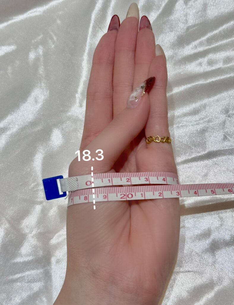 手鐲配戴測量教學：將大拇指往小指方向內縮，使用軟尺繞過手掌骨頭最寬處一圈，顯示周長約 18.3 公分。
